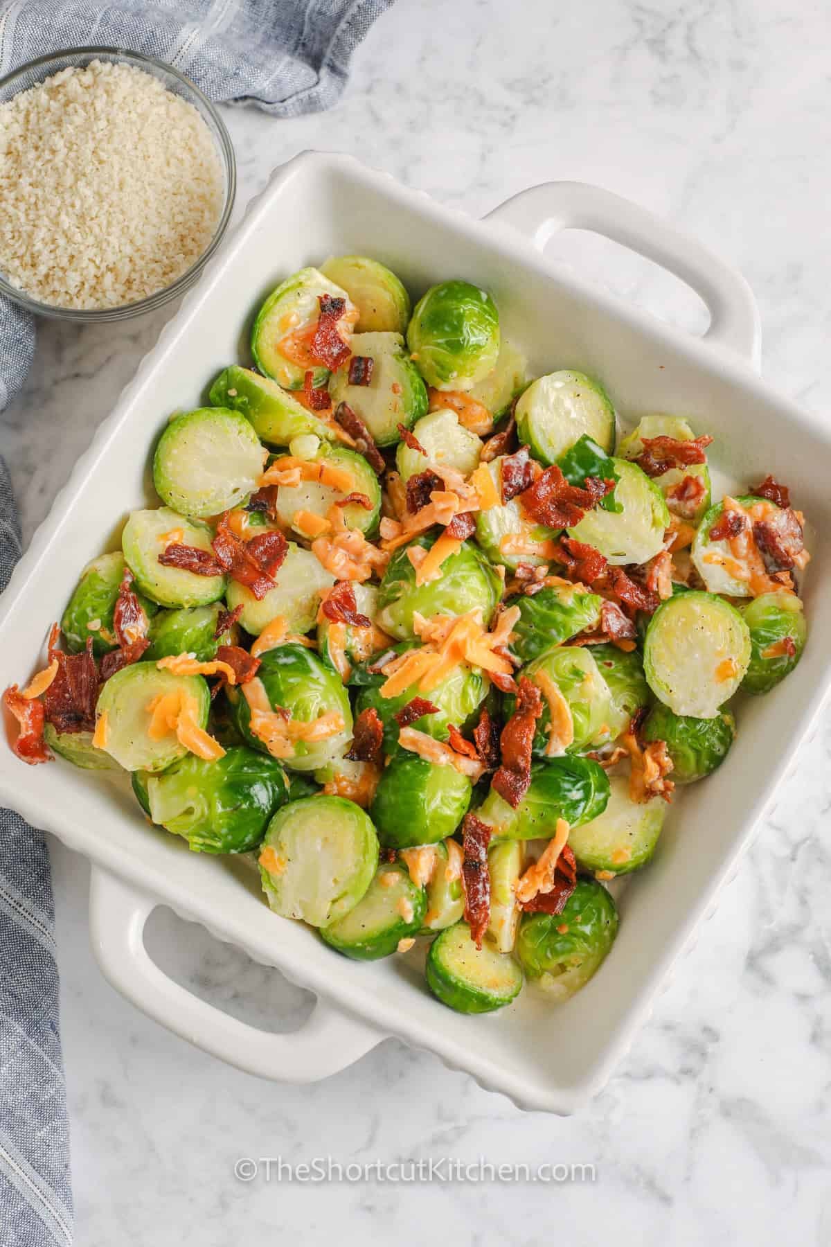Creamy Brussel sprout casserole gives an everyday vegetable a cozy upgrade with bacon, cheddar cheese, and crunchy panko. Halved Brussels sprouts cook quickly before being folded into rich cream and cheese and baked until the topping is golden. This easy baked Brussels sprouts recipe takes only 30 minutes from start to finish. Add this simple cheesy casserole to your weeknight or holiday menu. #theshortcutkitchen adding Brussels sprouts , cheese , bacon together in dish to make Brussel Sprout Casserole.