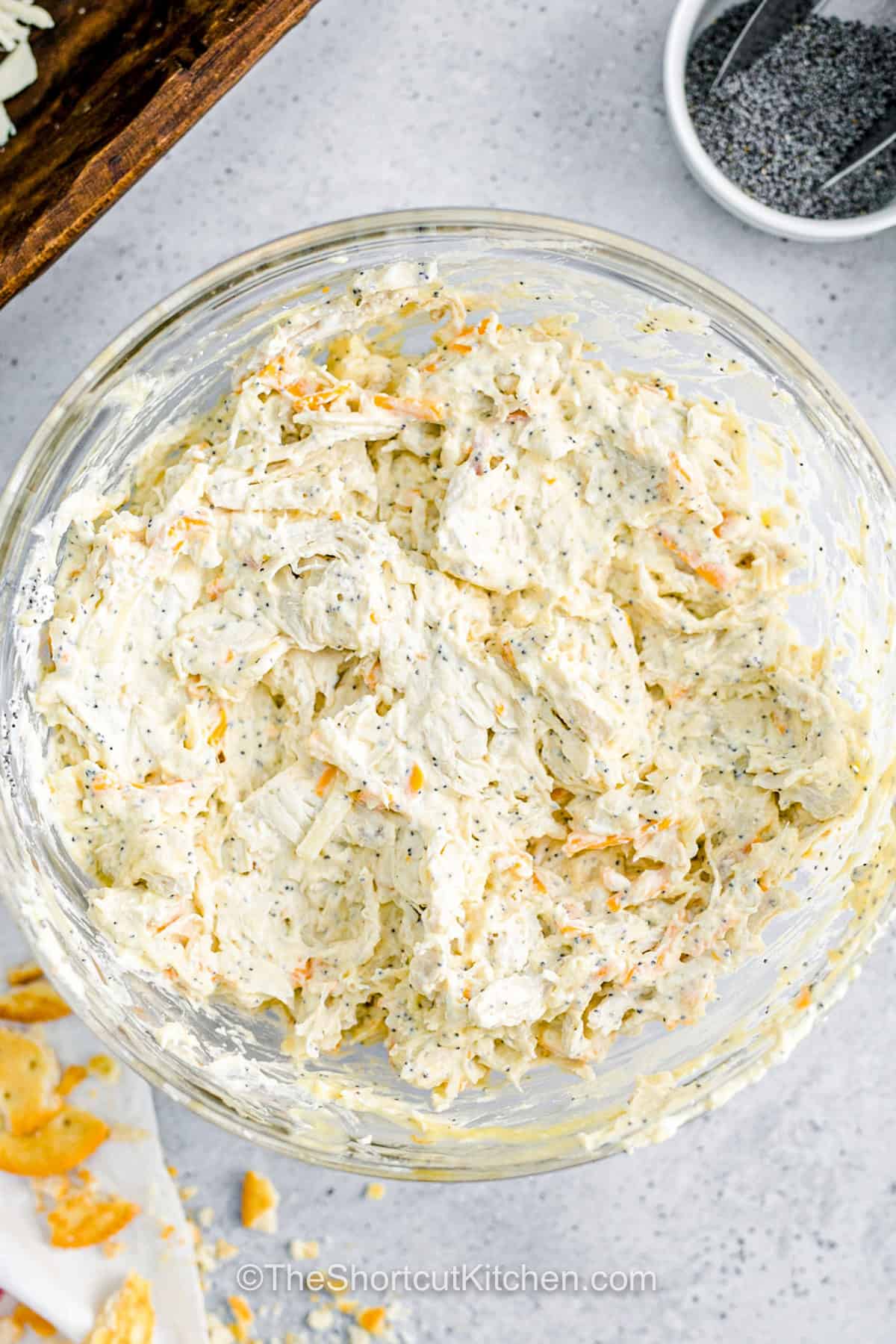 This creamy chicken poppyseed casserole has everything a cozy dinner needs, including tender chicken, melted cheese, a savory cream sauce, poppy seeds, and a golden Ritz cracker topping. The easy oven casserole takes just 5 minutes to assemble because the chicken is already cooked. Add optional bacon for smoky flavor or keep it classic. Save this chicken casserole with Ritz crackers for an easy family dinner. #theshortcutkitchen cheese and chicken mixing a bowl to make Chicken Poppyseed Casserole.