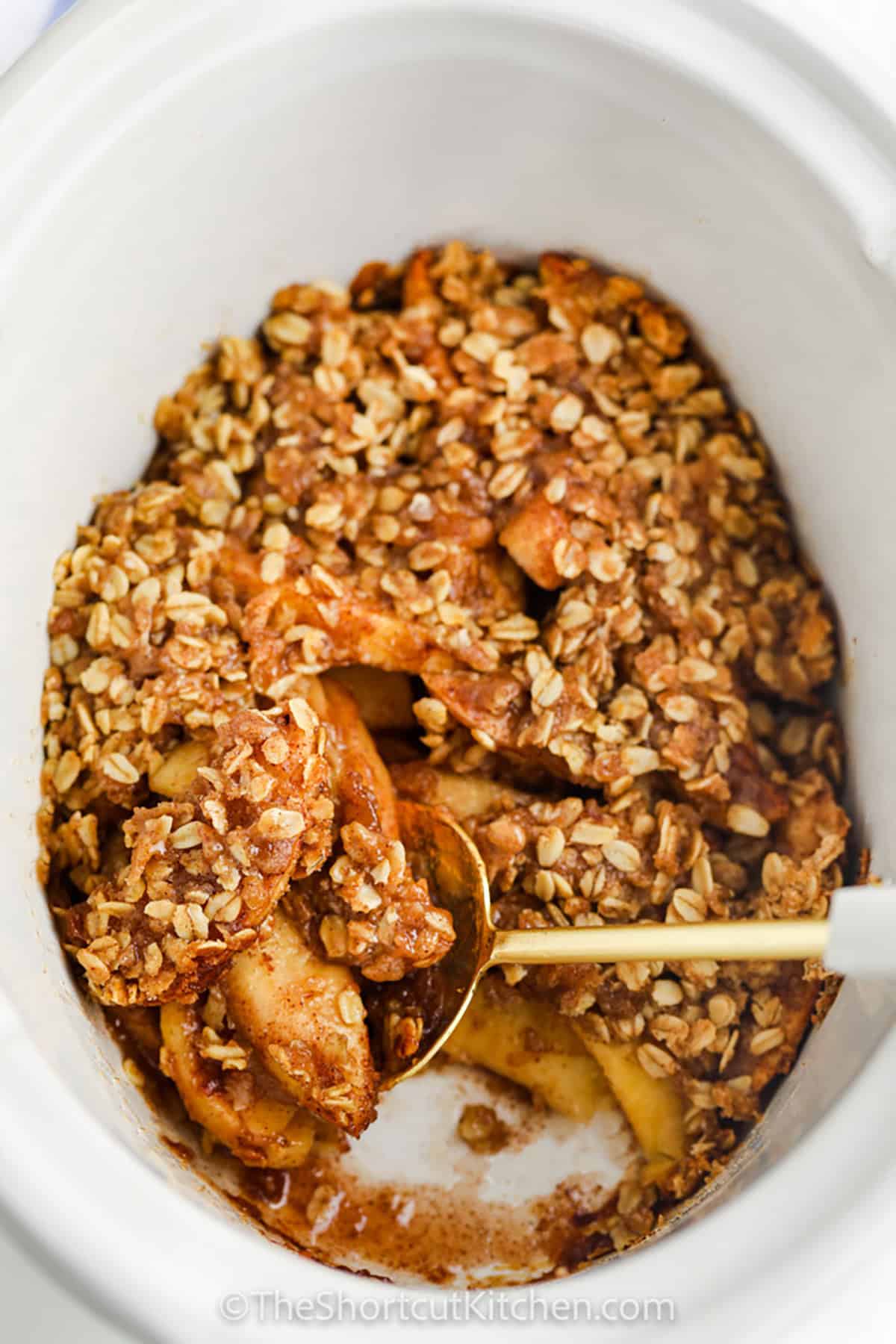 Slow Cooker apple crisp makes homemade fall dessert easy with fresh apples, cinnamon, brown sugar, and old-fashioned oats. The apples cook until tender beneath a buttery crumble topping, all without turning on the oven. This cozy crockpot apple dessert needs only 15 minutes of prep and is delicious served warm with vanilla ice cream. Save this easy apple crisp recipe for fall and make it tonight. #theshortcutkitchen top view of Crock Pot Apple Crisp in the pot with a spoon