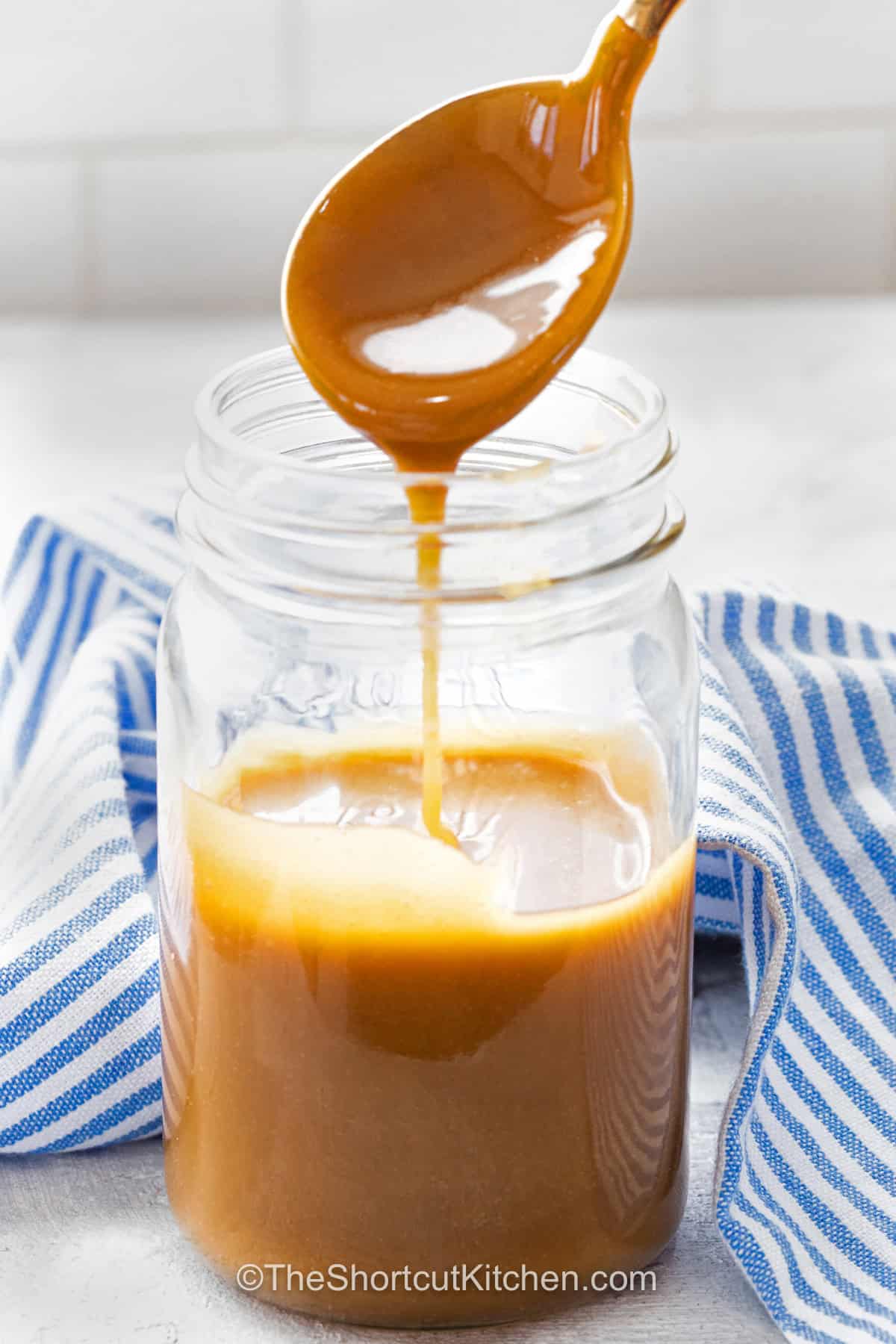 This easy caramel sauce turns butter, brown sugar, and milk into a rich homemade topping in minutes. The simple stovetop method makes a smooth brown sugar caramel sauce without the extra steps of traditional caramel. Drizzle it warm over vanilla ice cream, apple pie, pancakes, brownies, or cheesecake for an easy finishing touch. Save this 3 ingredient caramel sauce recipe for your next dessert. #theshortcutkitchen adding Easy Caramel Sauce to a jar with a spoon.