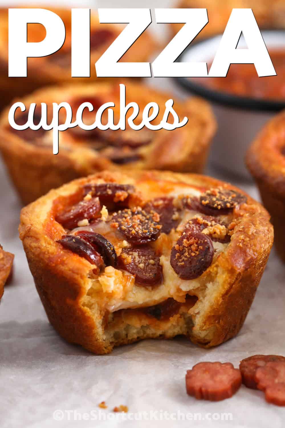 Pizza cupcakes are a fun twist on classic pizza, with flaky crescent roll edges and a cheesy center. Each little cup is filled with pizza sauce, mozzarella, and mini pepperoni for a snack everyone will enjoy. They are great for parties, game day, lunch boxes, or an easy family snack. Add your favorite pizza toppings to make them your own. Try these tasty pizza cupcakes for your next gathering. #theshortcutkitchen close up of Pizza Cupcakes with a bite taken out of one with a title.