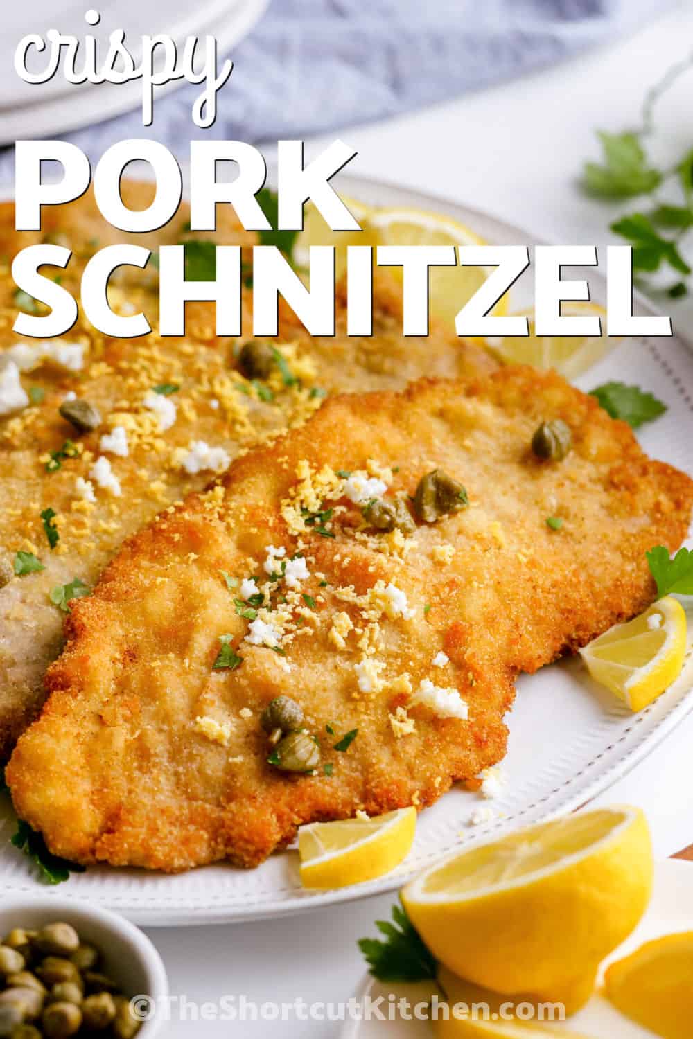 Looking for a simple way to cook pork tenderloin for dinner? This pork schnitzel recipe transforms it into thin breaded pork cutlets with a crisp golden exterior and tender center. Fine breadcrumbs make an even crunchy coating, while lemon, parsley, and capers add fresh flavor after frying. Pair this easy pork schnitzel with potatoes or cabbage for a satisfying meal. Pin this recipe for your dinner rotation. #theshortcutkitchen Pork Schnitzel Recipe with lemon and capers with a title.