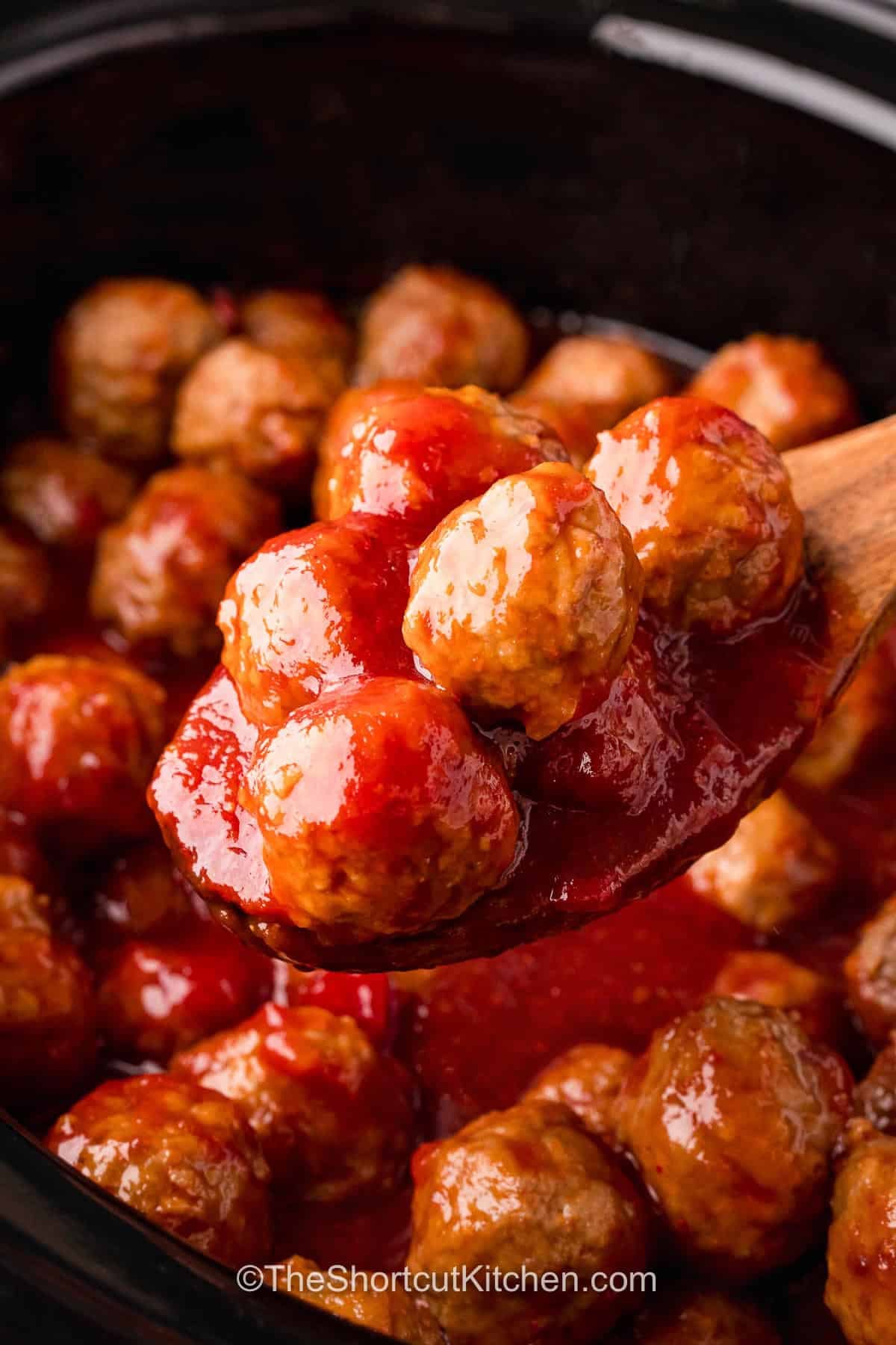Serve pepper jelly meatballs when you want an appetizer that is easy and full of flavor. The sweet raspberry jam balances the tangy chili sauce, while hot pepper jelly adds a pleasant kick. Tender meatballs simmer in the flavorful sauce until they are ready for serving. Keep them warm in the slow cooker and let guests grab a bite whenever they like. Make these easy crockpot meatballs for your next party and enjoy a delicious crowd-pleaser. #theshortcutkitchen Pepper Jelly Meatballs on a wooden spoon.