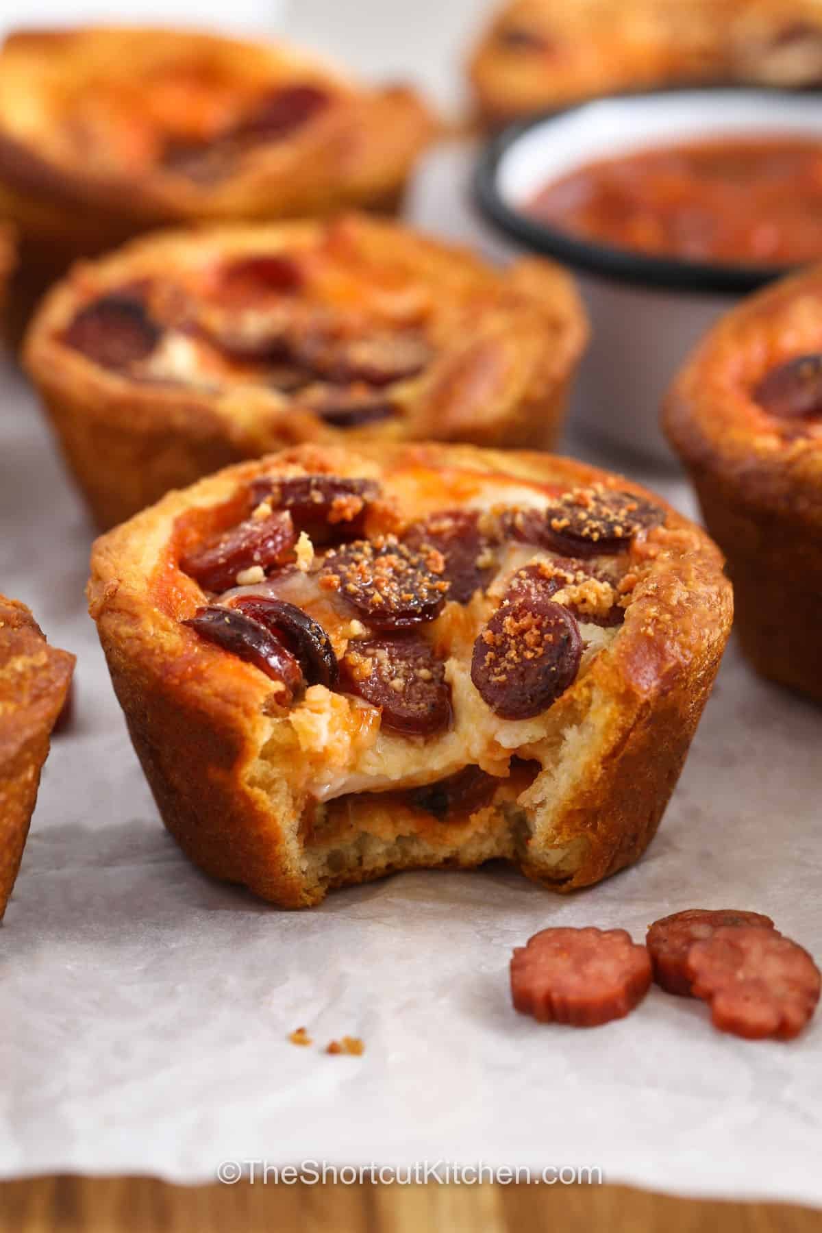 Easy pizza cupcakes are packed with the classic flavors everyone loves, all baked into cute individual portions. Crescent roll dough creates golden edges around pizza sauce, mozzarella, pepperoni, and Parmesan. They make a tasty appetizer for a party or a fun snack for the family. Add mushrooms, olives, sausage, or your favorite toppings for a personalized bite. Bake these pizza cupcakes for your next snack. #theshortcutkitchen Pizza Cupcakes with a bite taken out of one.
