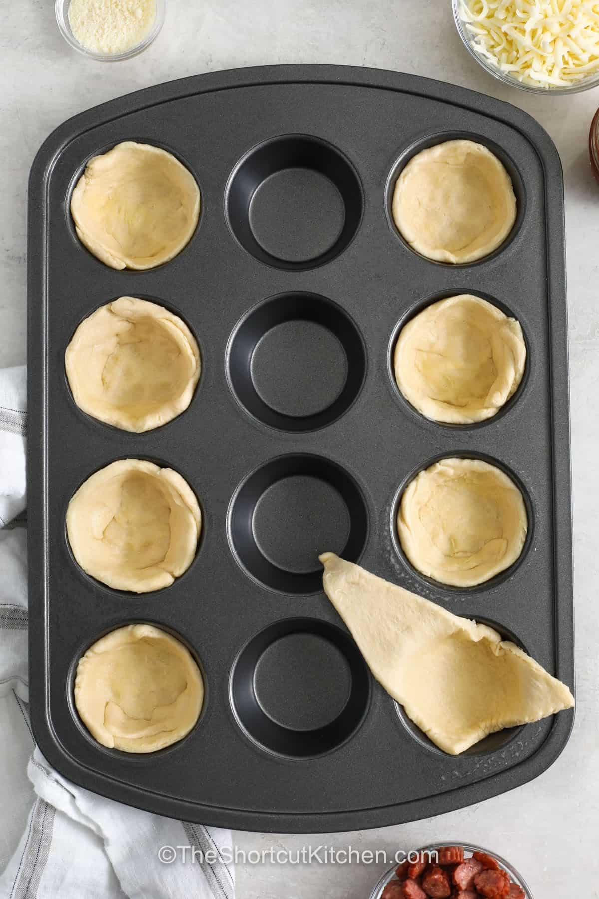 These pizza cupcakes turn simple crescent roll dough into a fun handheld pizza snack. Pizza sauce, mozzarella, pepperoni, and Parmesan come together in each golden cup for plenty of classic flavor. They are easy to customize with your favorite toppings and make a tasty appetizer, lunch, or after-school snack. Keep this easy muffin tin recipe handy for busy days. Give these pizza cupcakes a try today. #theshortcutkitchen adding crescent roll dough to muffin tins to make Pizza Cupcakes.