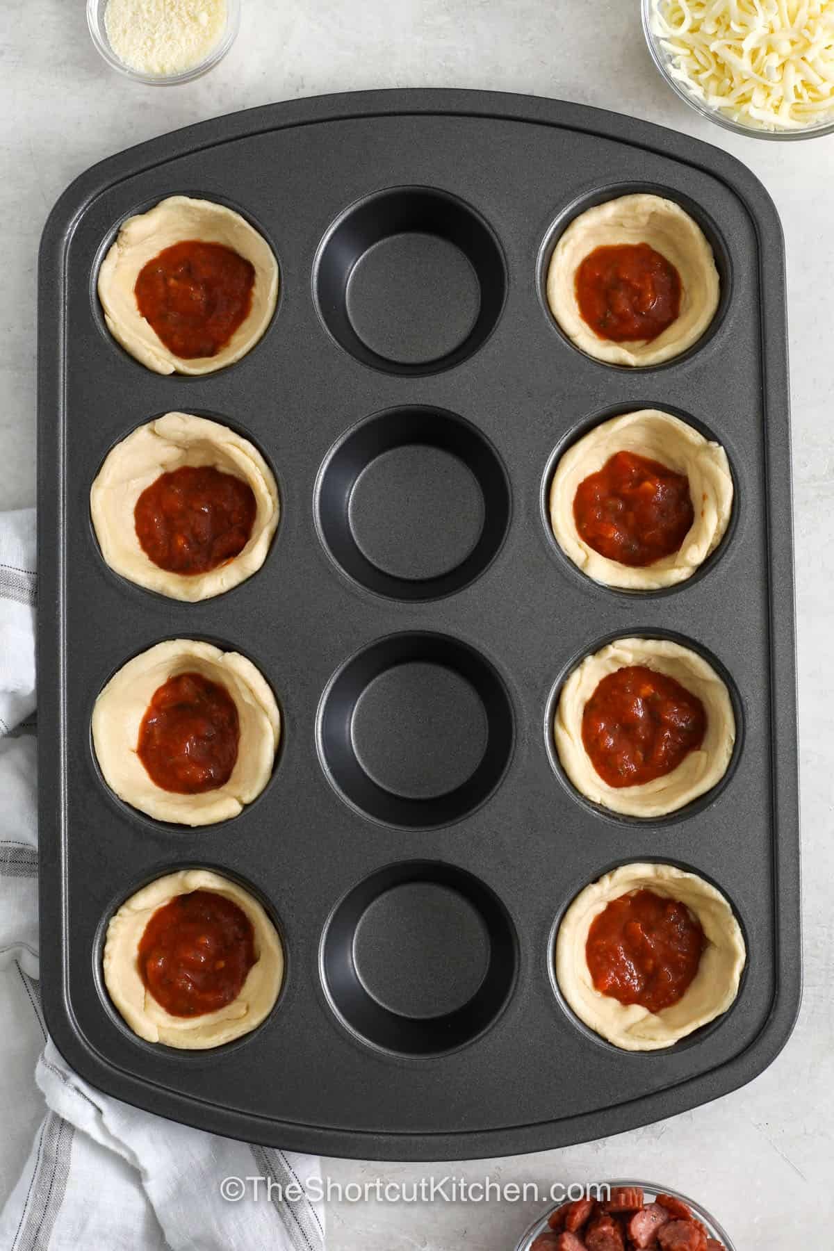 Pizza cupcakes are a fun twist on classic pizza, with flaky crescent roll edges and a cheesy center. Each little cup is filled with pizza sauce, mozzarella, and mini pepperoni for a snack everyone will enjoy. They are great for parties, game day, lunch boxes, or an easy family snack. Add your favorite pizza toppings to make them your own. Try these tasty pizza cupcakes for your next gathering. #theshortcutkitchen adding pizza sauce to crescent doll roll to make Pizza Cupcakes.