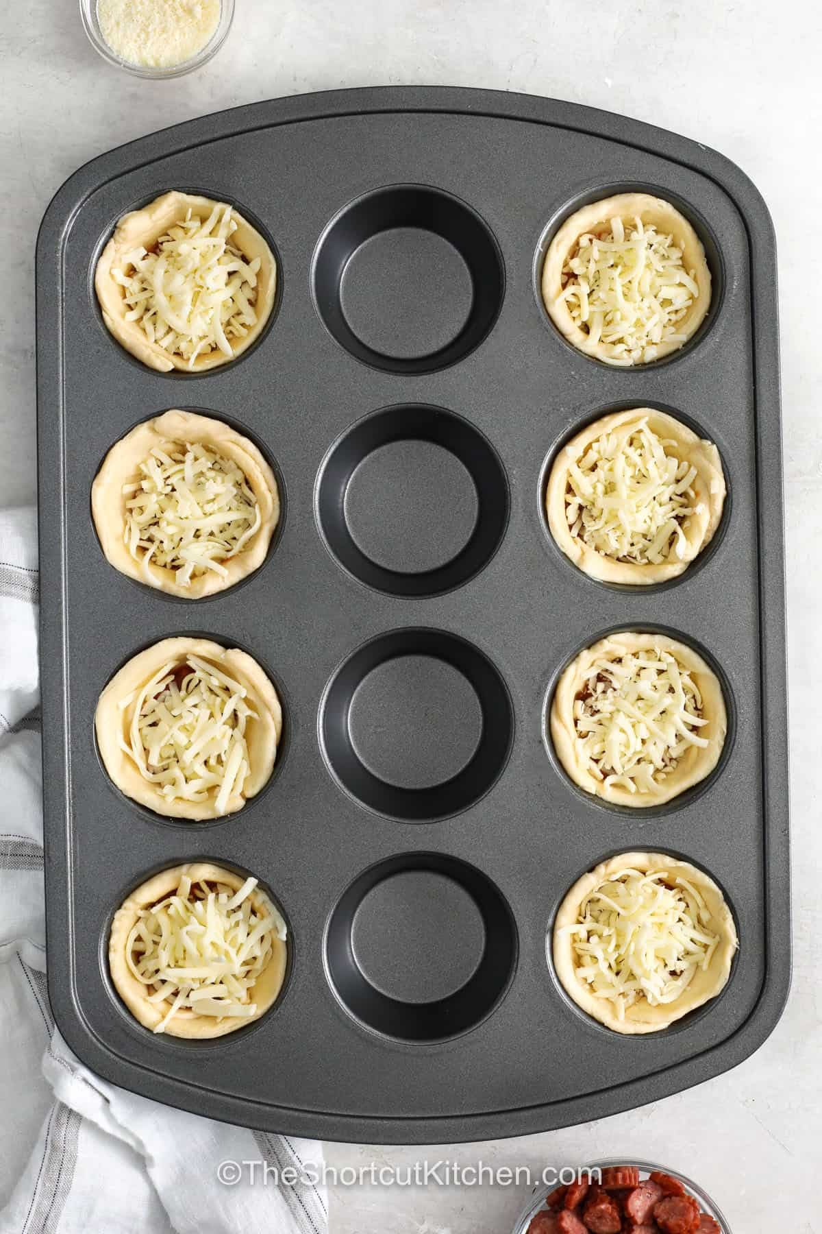 Pizza cupcakes are a tasty way to turn everyday ingredients into a fun family favorite. Crescent roll dough bakes around pizza sauce, mozzarella, and pepperoni for a cheesy center and golden edges. They work well as an easy appetizer, snack, or casual lunch, and you can add any pizza toppings your family enjoys. Keep this simple muffin tin recipe ready for busy days. Try these pizza cupcakes today. #theshortcutkitchen adding cheese on top of Pizza Cupcakes.