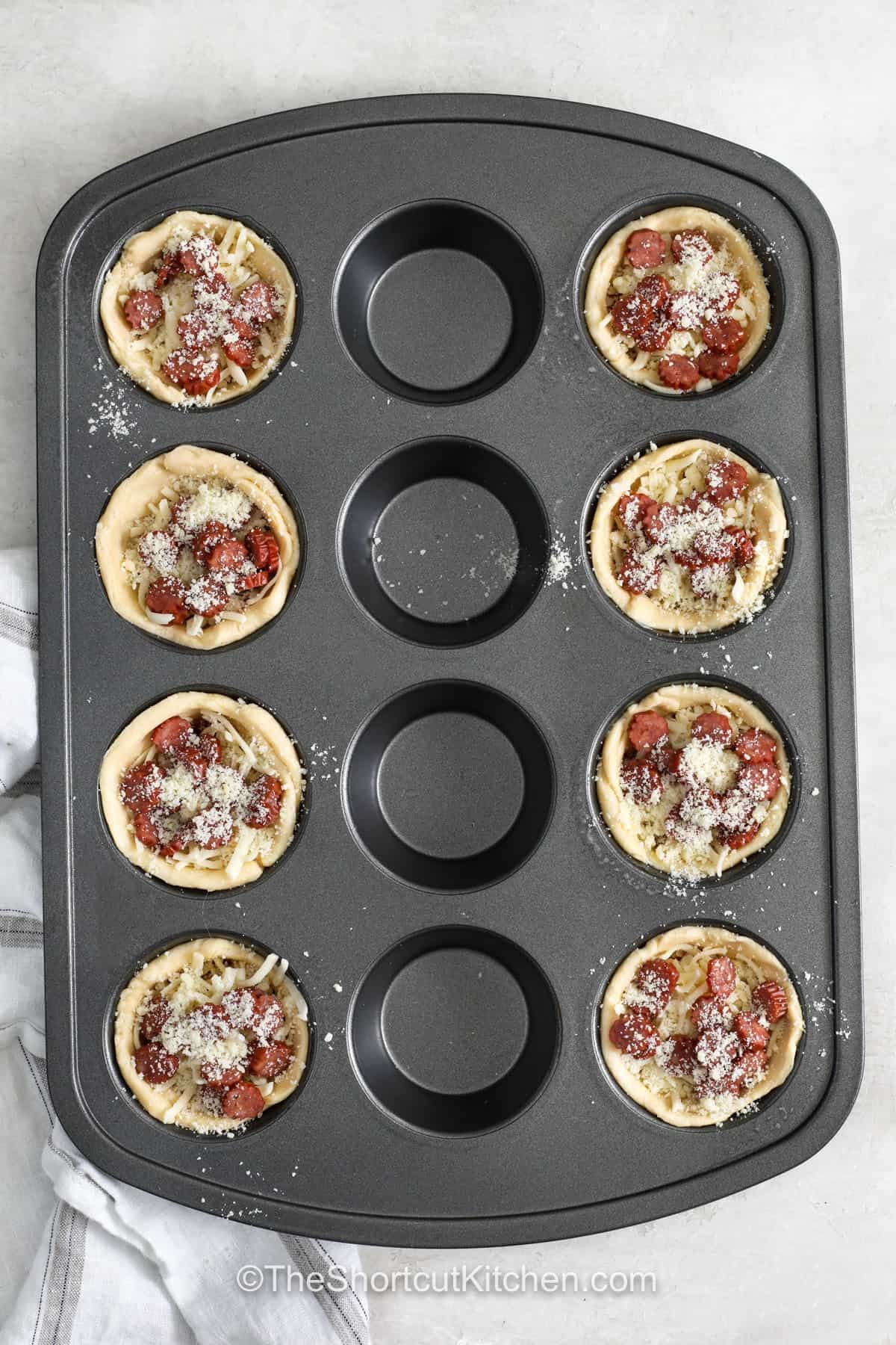 For an easy party snack, pizza cupcakes are hard to resist. Crescent roll dough forms little cups filled with pizza sauce, shredded mozzarella, and mini pepperoni, then each one gets a sprinkle of Parmesan. They are fun to serve, easy to customize, and full of familiar pizza flavor. Try different toppings for a new version every time. Make these cheesy pizza cupcakes for your next gathering. #theshortcutkitchen adding pepperoni and parmesan to Pizza Cupcakes before cooking.