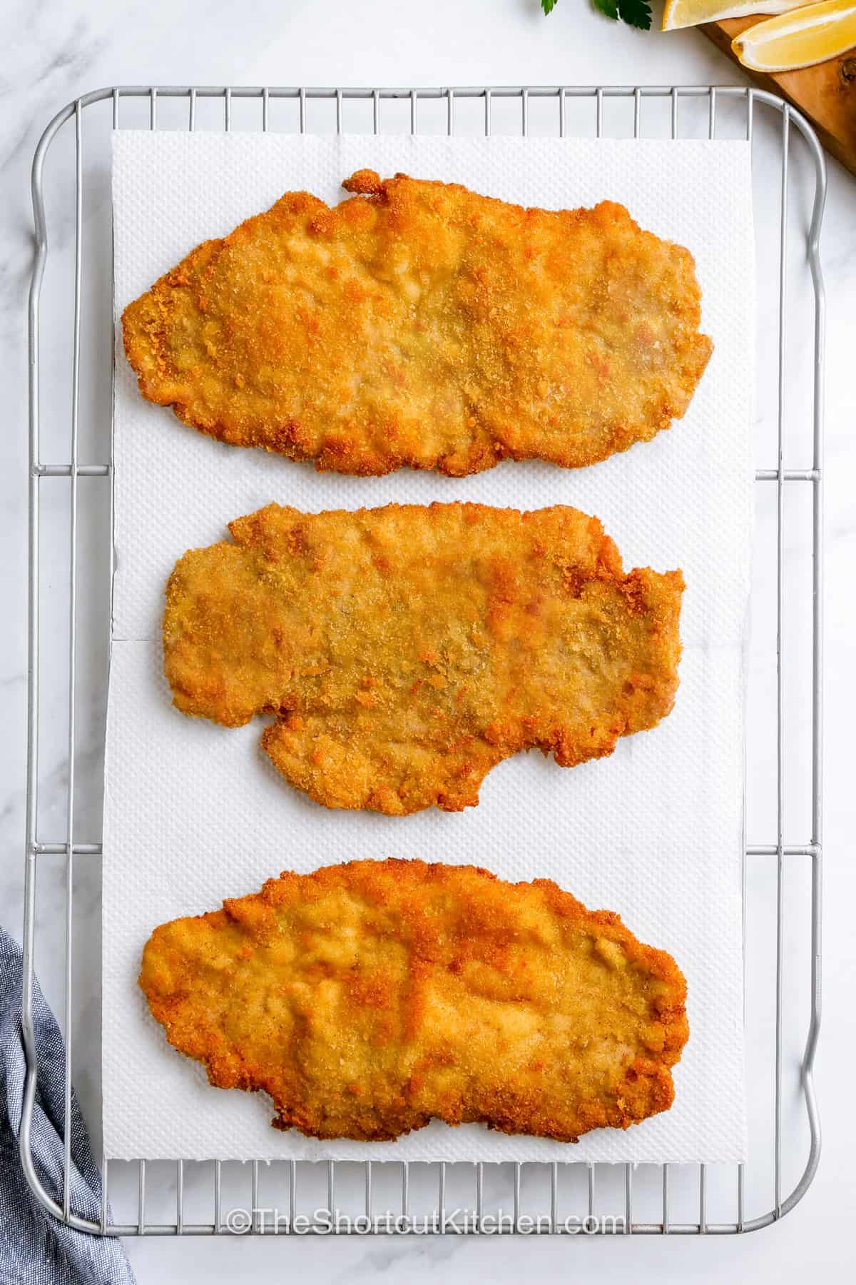 This easy pork schnitzel recipe delivers everything a good breaded pork cutlet should have, a crunchy golden crust, tender pork tenderloin, and plenty of fresh lemon. Pounding the pork thin helps it cook quickly, while a flour, egg, and breadcrumb coating creates irresistible texture. Finish the pan fried pork schnitzel with parsley and capers. Pin it for an easy family dinner everyone will love. #theshortcutkitchen Pork Schnitzel Recipe on paper towel and cooling rack.