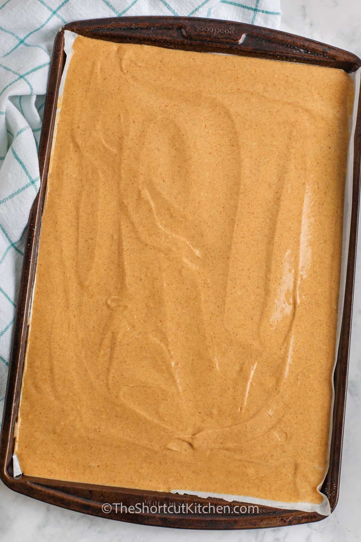 Cake mix makes this pumpkin roll cake surprisingly simple. Pumpkin puree and pumpkin pie spice give the thin baked cake plenty of warm fall flavor before it is wrapped around a fluffy cream cheese filling. Chill the finished pumpkin cake roll for neat slices and an easy make-ahead dessert. It is a festive shortcut recipe for Thanksgiving and fall parties. Pin this cake recipe for later. #theshortcutkitchen adding Pumpkin Roll Cake to the sheet pan.