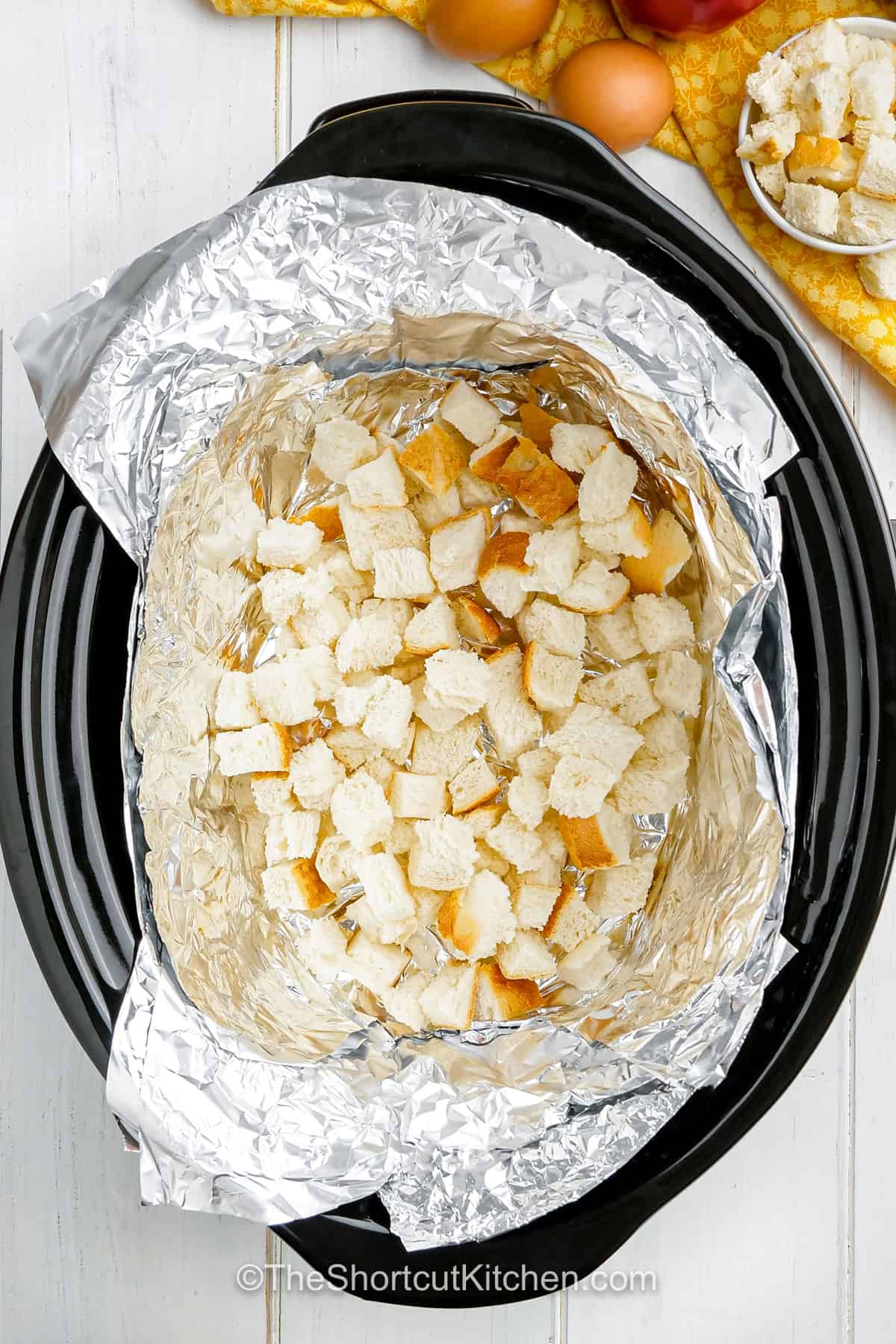 Make holiday breakfast easier with Slow Cooker breakfast strata casserole. The bread, vegetables, and chorizo can be prepared ahead, then layered with cheese and egg custard in the slow cooker. A foil collar helps protect the edges from overcooking, while a foil sling makes the finished strata easy to lift out. It’s a simple, hands-off way to feed a crowd. Make this recipe for breakfast today. #theshortcutkitchen bread cubes in the bottom of the slow cooker in the slow cooker breakfast strata casserole.