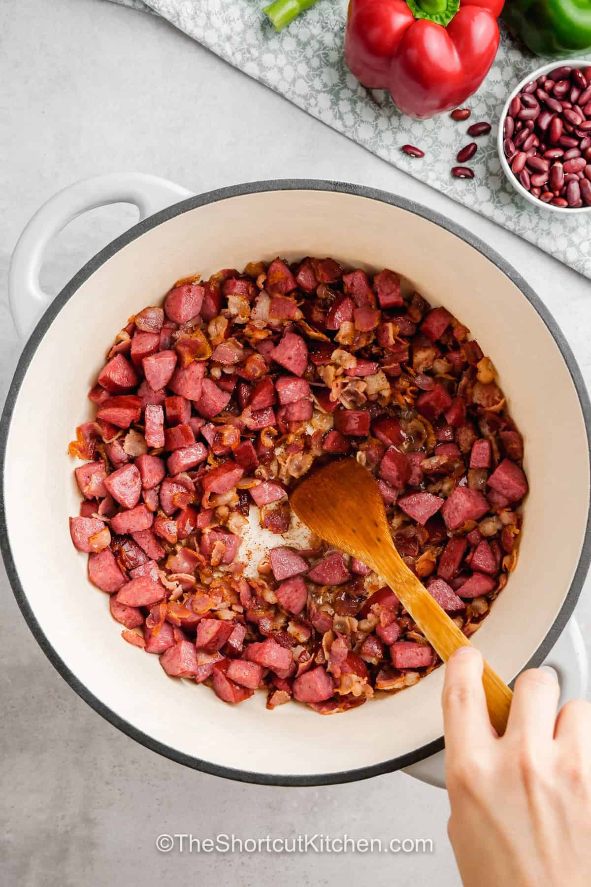 A big pot of easy red beans and rice makes comfort food simple. Dried kidney beans simmer with andouille sausage, smoky bacon, peppers, celery, bay leaves, thyme, cayenne, and chicken broth before being served over warm white rice. This stovetop red beans and rice with sausage recipe is hearty, flavorful, budget-friendly, and great for leftovers. Pin it now for an easy family dinner. #theshortcutkitchen cooking sausage and onion in pan to make Easy Red Beans and Rice .