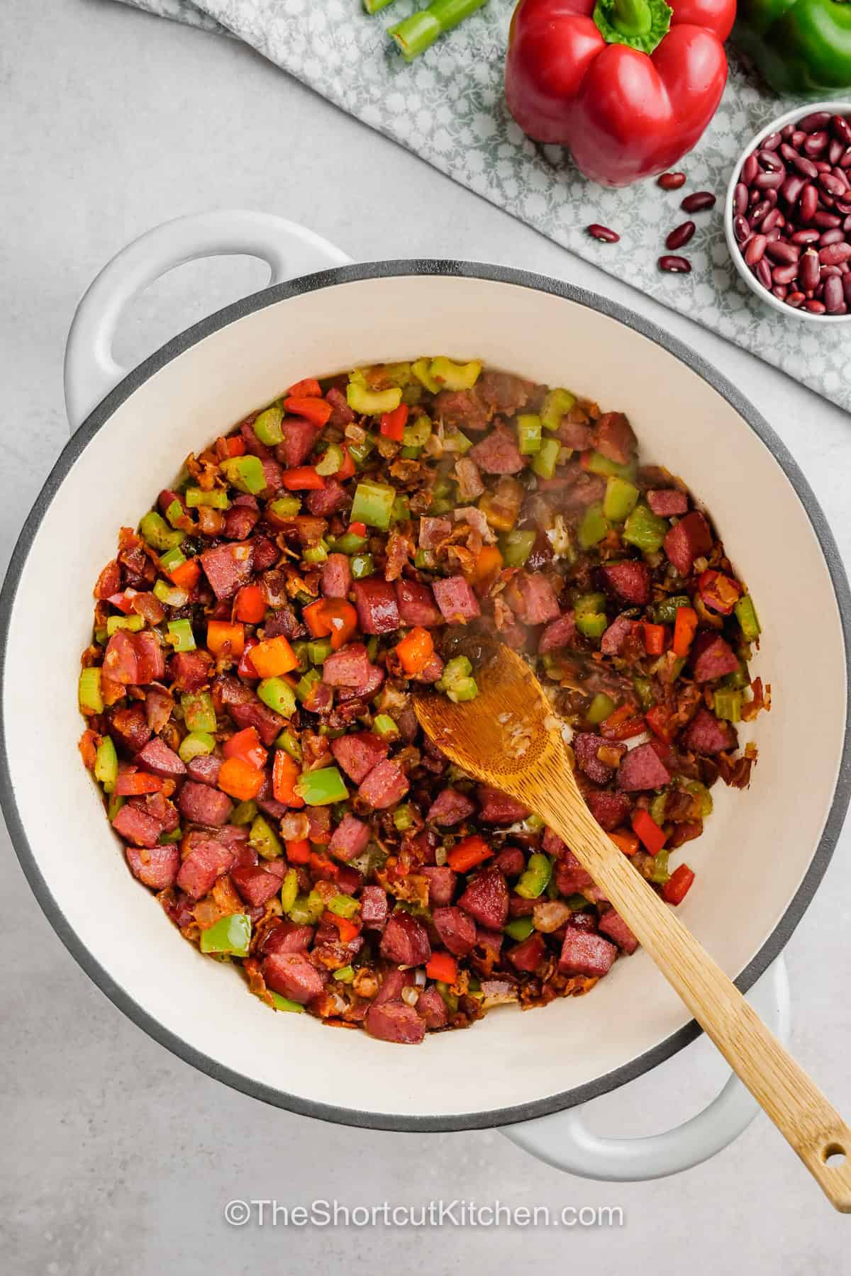 For a hearty family dinner, make this easy red beans and rice recipe with dried kidney beans and smoky andouille sausage. Bacon, peppers, celery, herbs, and chicken broth add layers of flavor while a slow stovetop simmer gives the beans their tender texture. Serve the finished red beans and rice with green onions and extra hot sauce. Save this easy comfort food recipe for your next meal plan. #theshortcutkitchen adding peppers and celery to pot of sausage and onions to make Easy Red Beans and Rice